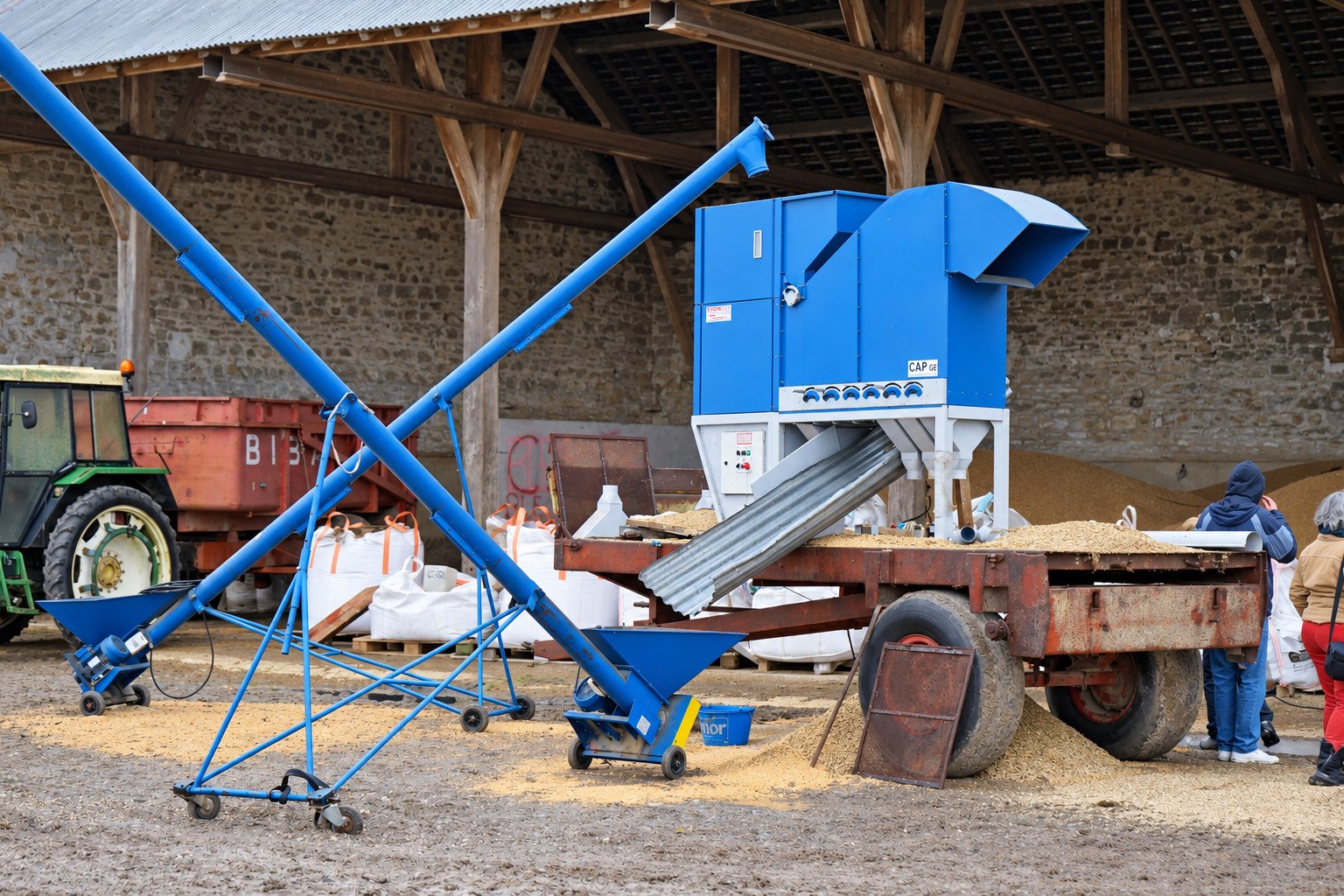 Le trieur bleu et sa vis d'alimentation installés dans une cour de ferme, un tracteur derrière