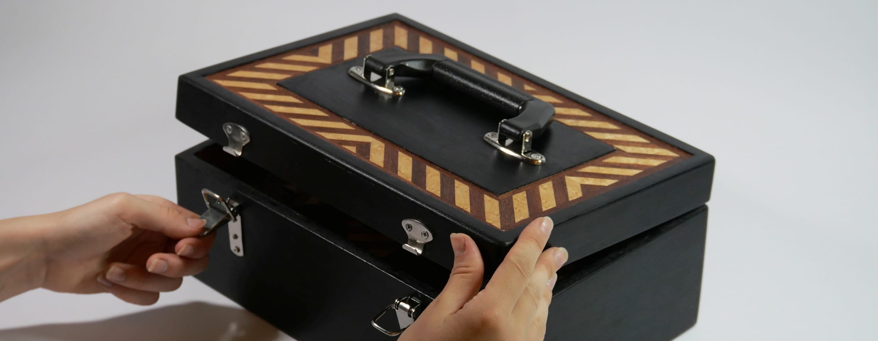 Deux mains ouvrent le coffret noir en bois, son couvercle bordé d'un motif de marqueterie en chevrons